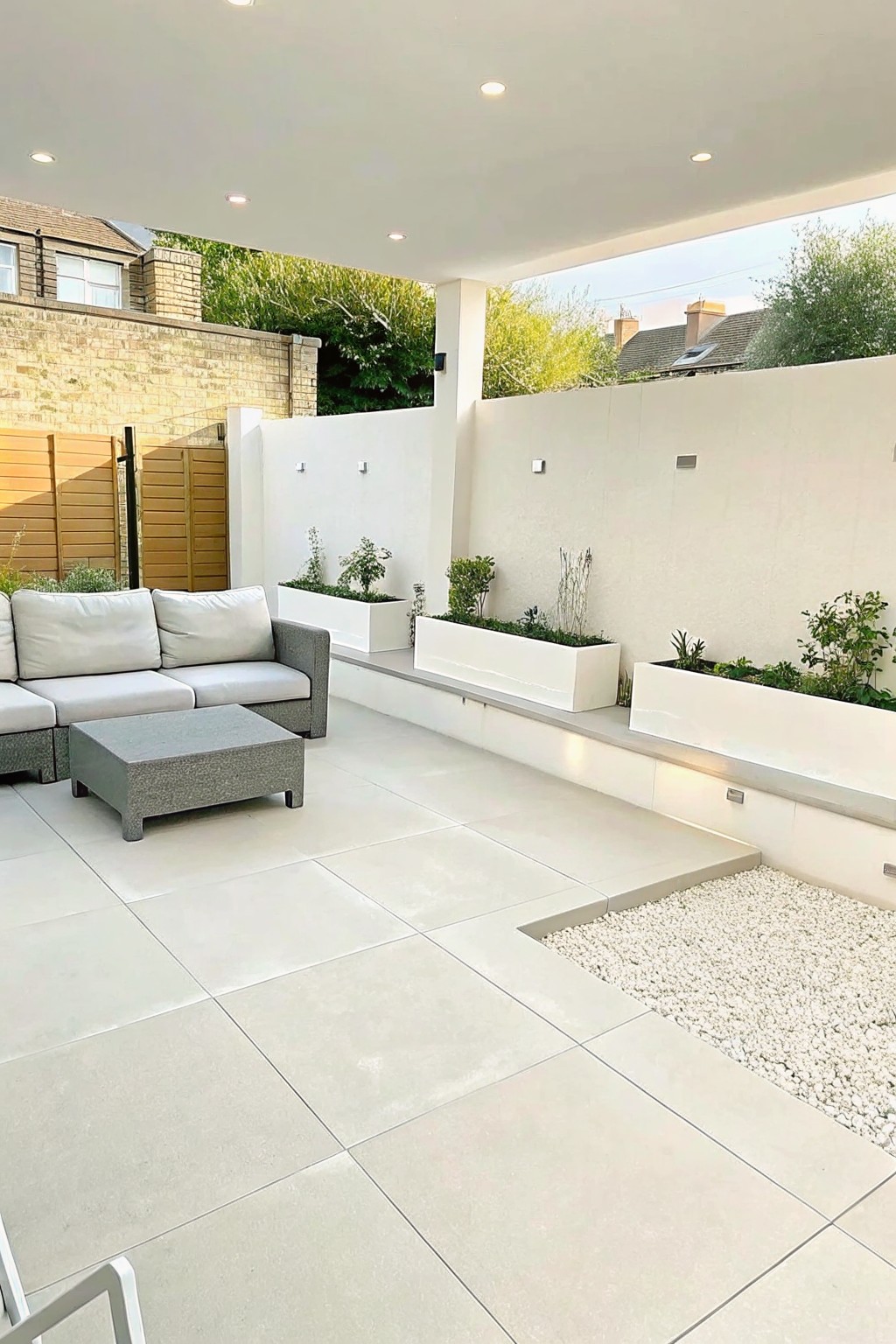 An outdoor patio with large light gray tiles, a gray sectional sofa and ottoman, and multiple long white rectangular planters arranged along white walls with built-in lighting.