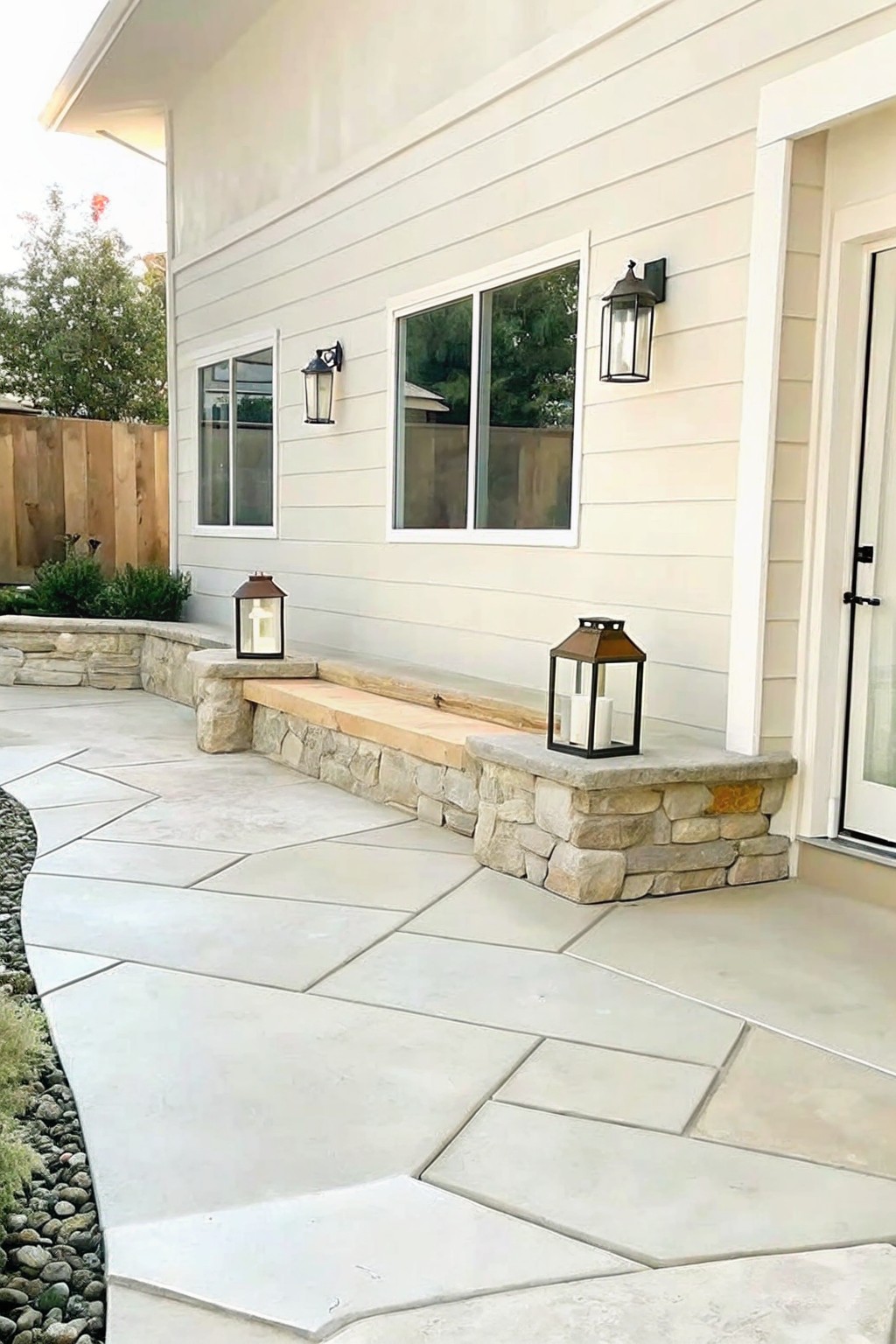 A house exterior with light siding shows a patio of irregular gray stone pavers and a long bench made from a wooden beam resting on stacked natural stone walls, with two lanterns placed on the stone.
