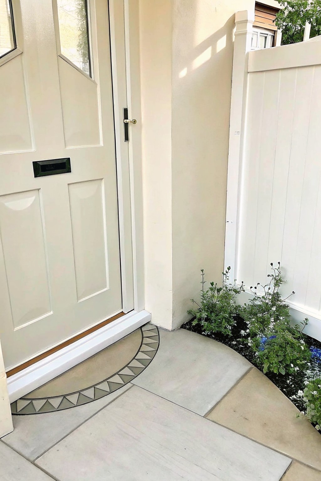White door with doormat on paved path beside white fence and garden bed.