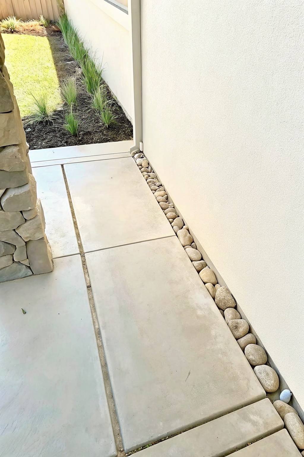 A concrete patio with large pavers next to a white wall, edged with a row of smooth river rocks.