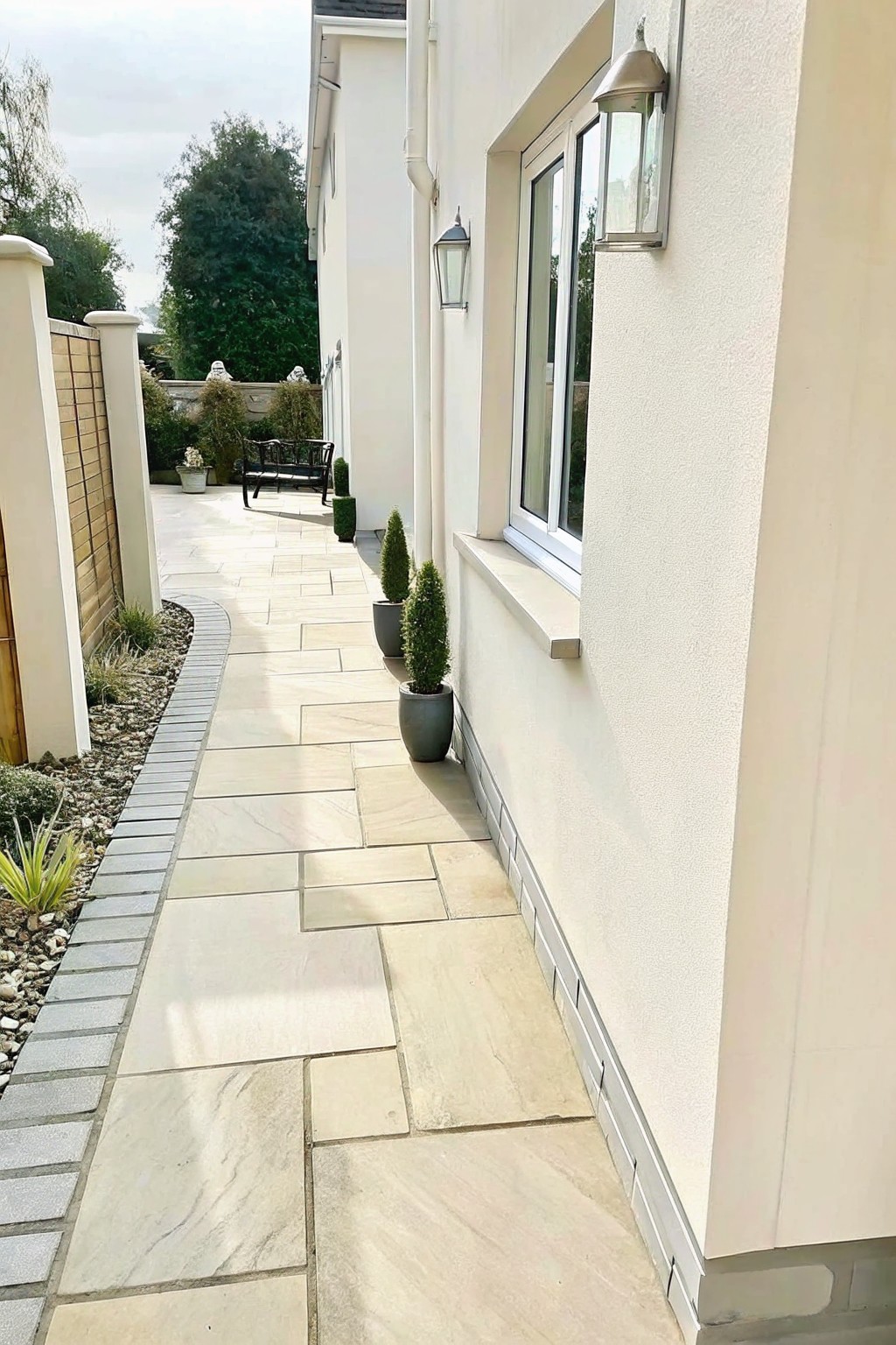 A long pathway of irregular light-colored stone pavers runs alongside a white house wall, bordered by a gravel bed with plants on the left and small potted shrubs on the right.