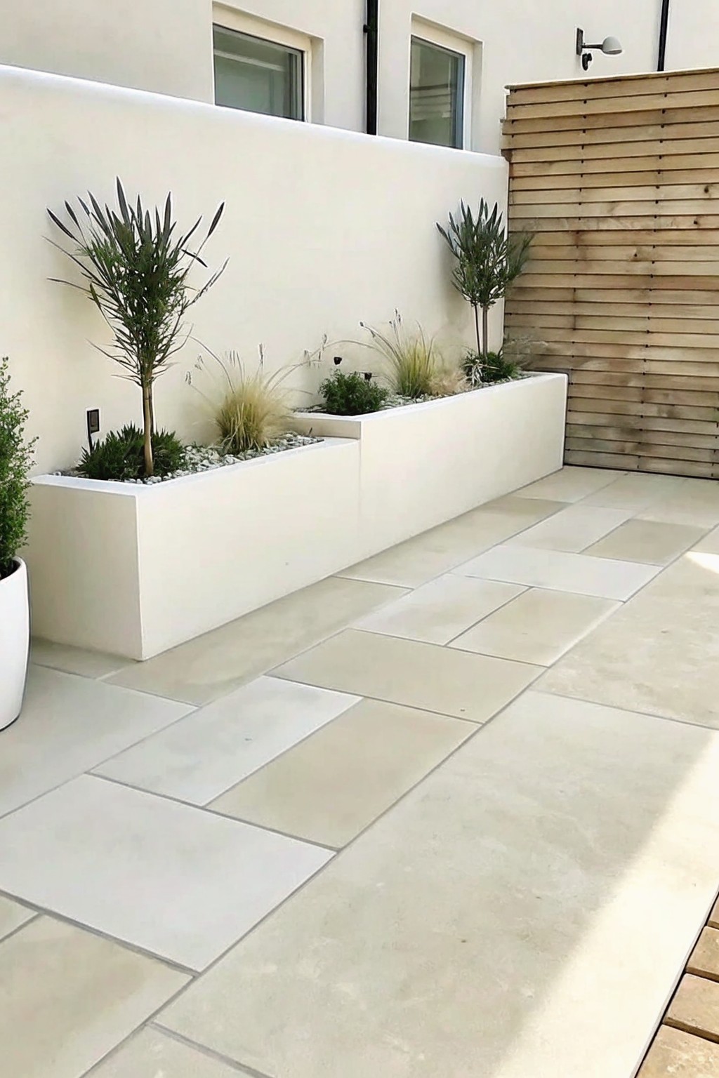 Light stone patio tiles beside white raised planters holding grasses and small shrubs against a plain wall.