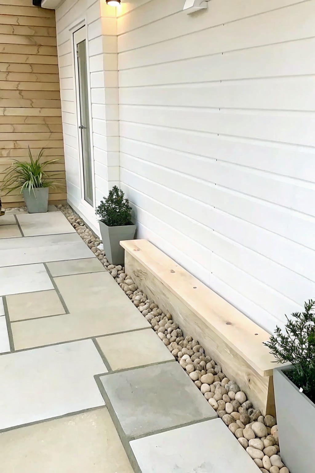 A wooden timber beam borders a strip of river rock along the side of a white house next to large stone pavers.