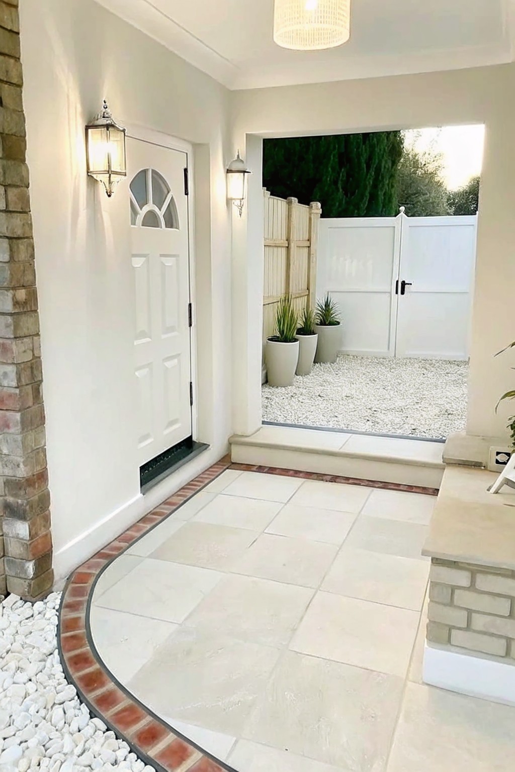 A white front door opens onto a tiled porch with curved brick edging that separates the tiles from a white gravel area containing potted plants and a white gate.