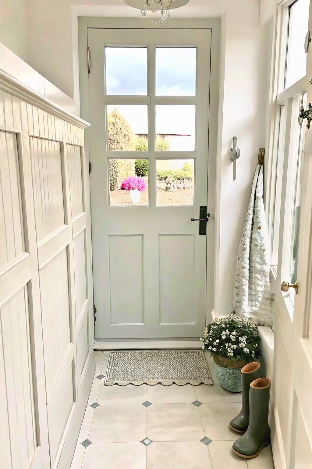 Sage green door with glass panes in white entryway beside green boots and flowers