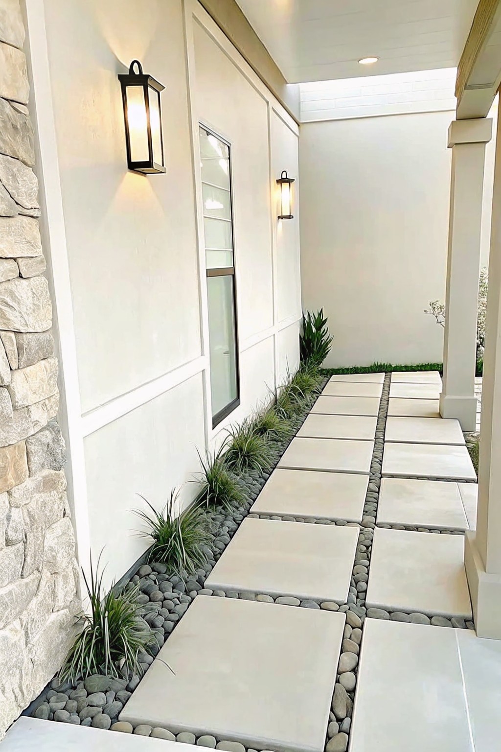 A stone paver walkway runs beside a white house wall, bordered by gray gravel and clumps of ornamental grass.