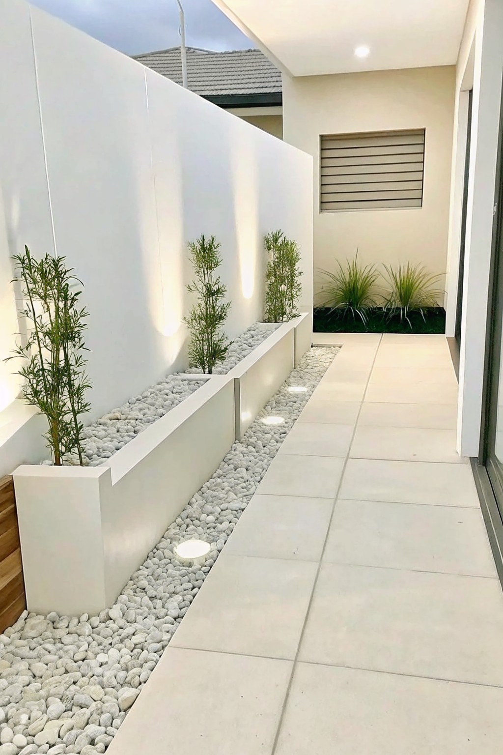A narrow paved walkway bordered by long white planters filled with white pebbles, small trees, and grasses, set against white walls.