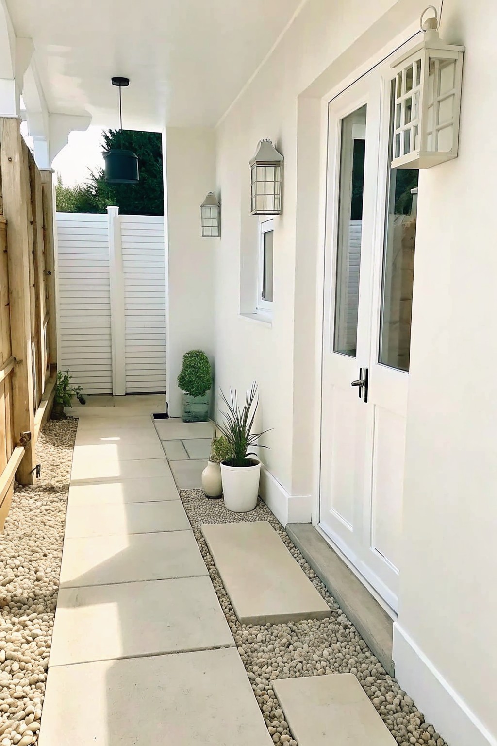 A narrow outdoor walkway with rectangular concrete pavers set into gravel, running beside a white building wall with doors and wall lanterns.