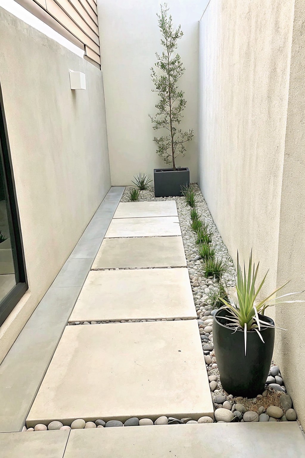 A narrow outdoor walkway lined with large rectangular concrete stepping stones set in white gravel, bordered by pebbles and potted plants between two walls.