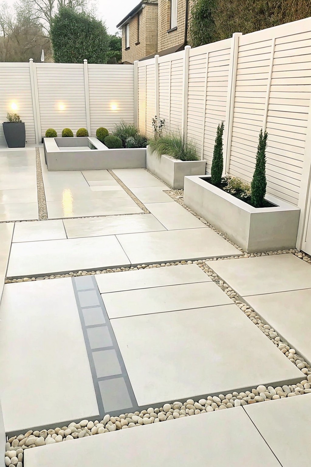 A modern garden patio with large light-colored paving slabs separated by bands of small white pebbles and bordered by concrete raised planters.