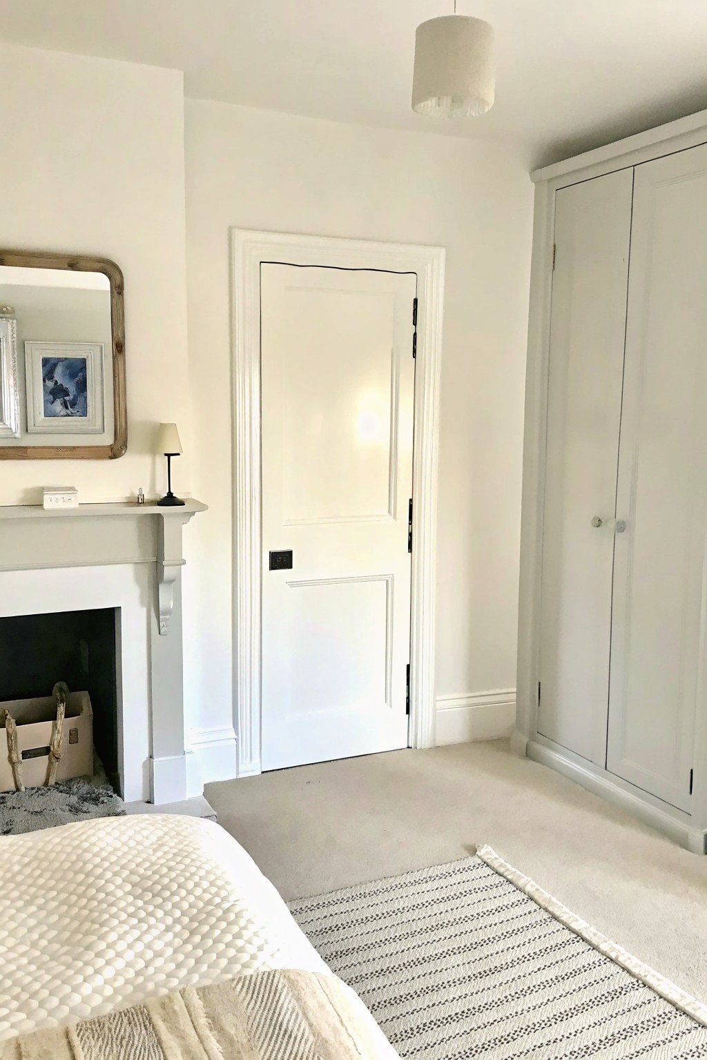 A bedroom interior with a white paneled door, light carpet, and a striped rug with a visible border placed near the foot of a bed.