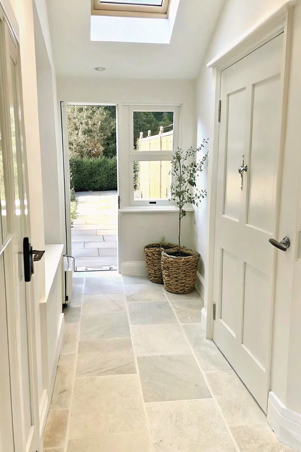 Light stone tile hallway leading to an open door with a matching stone path outside bordered by shrubs.