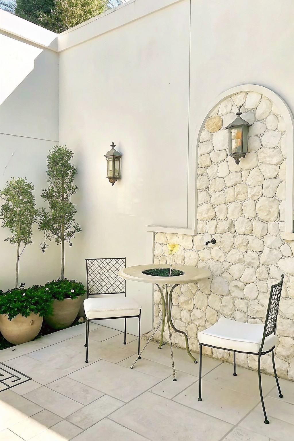 An outdoor patio with large light-colored tiles that include a black geometric border pattern along one edge, two black metal chairs with white cushions, a round table, and several large potted plants against a white wall.