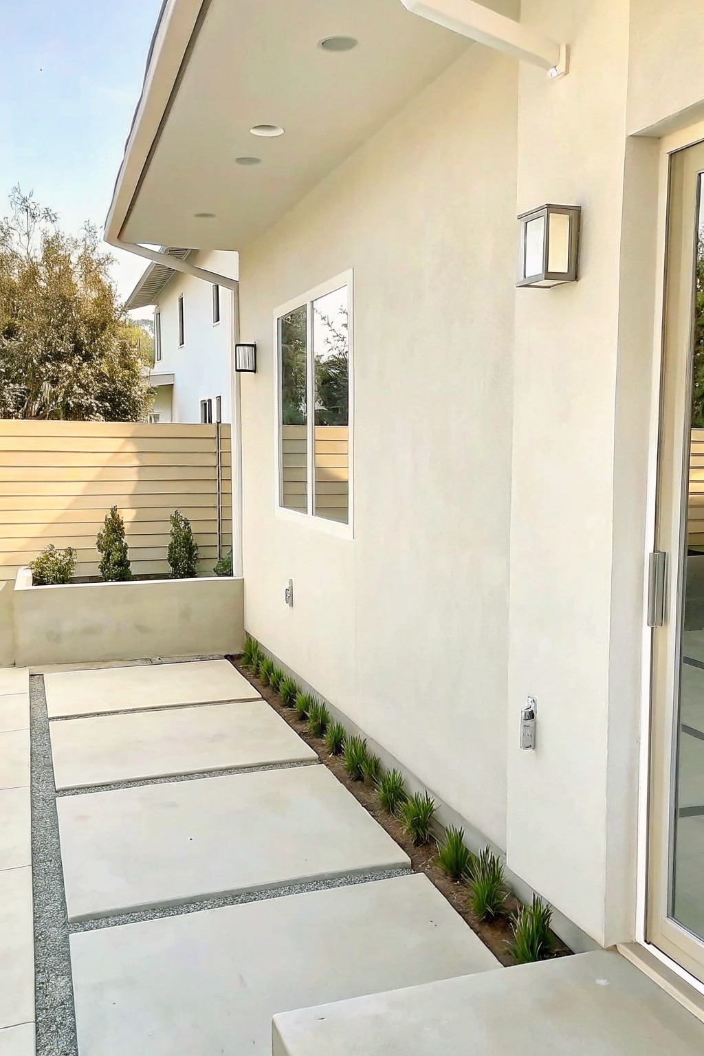 Modern beige house side with concrete pavers, plants, window, door, and wall lights