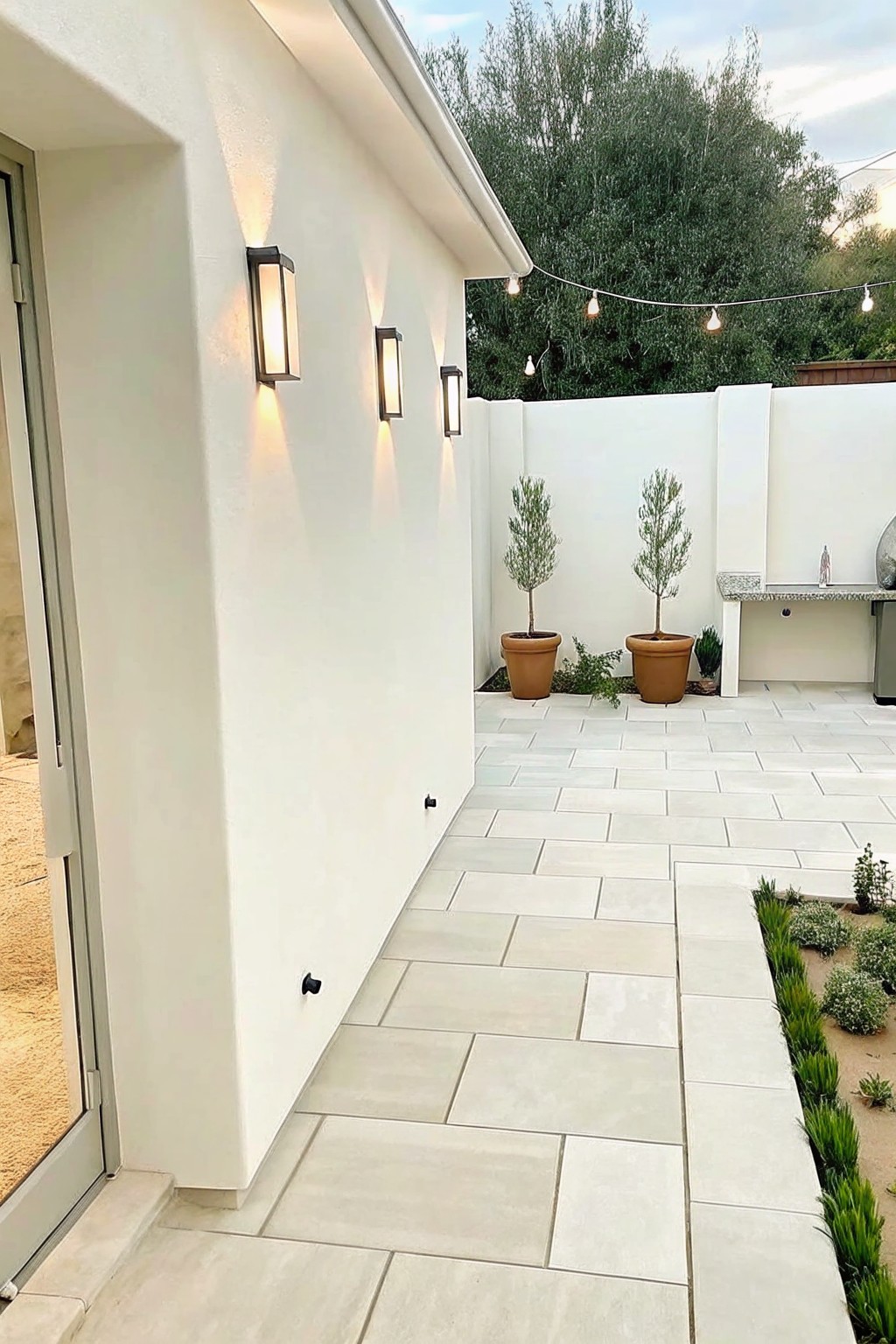 Modern white patio with glowing wall sconces, potted trees, and string lights