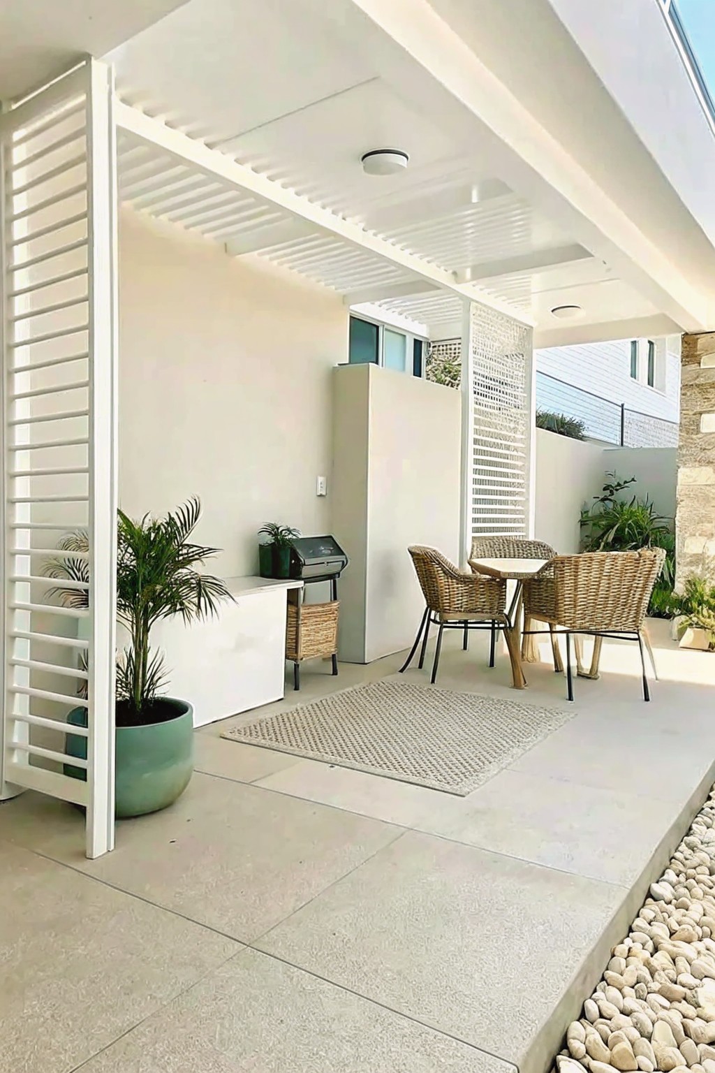 An outdoor concrete patio with a large teal pot holding a palm plant, wicker chairs around a small table, a woven rug, and a white slatted screen on the left.
