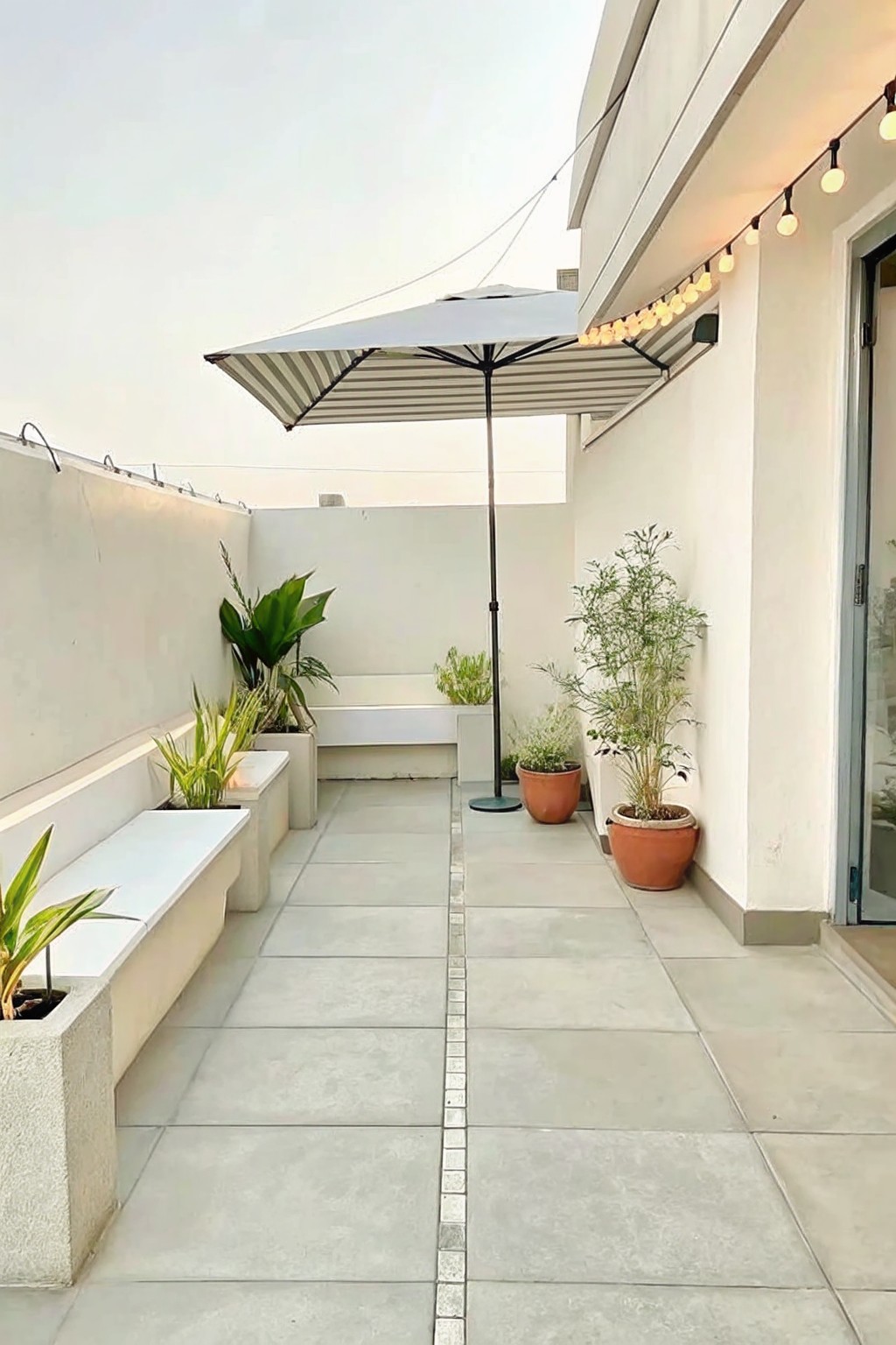 Modern rooftop patio with striped umbrella, string lights, potted plants, and tiled floor