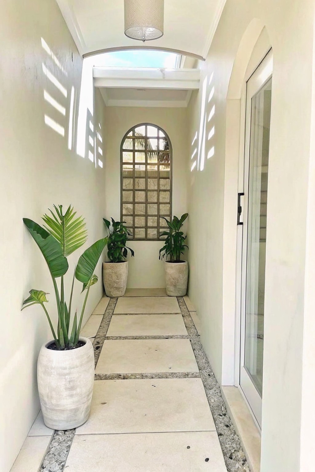 A narrow white interior hallway with large potted tropical plants along one wall, stone tile flooring, an arched window at the end, and a glass door on the right.