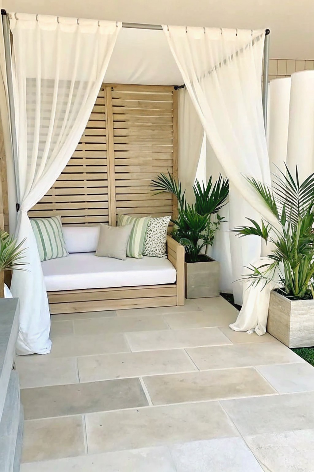 An outdoor wooden daybed with white curtains sits on a stone patio, flanked by two large rectangular planters holding tall tropical plants.