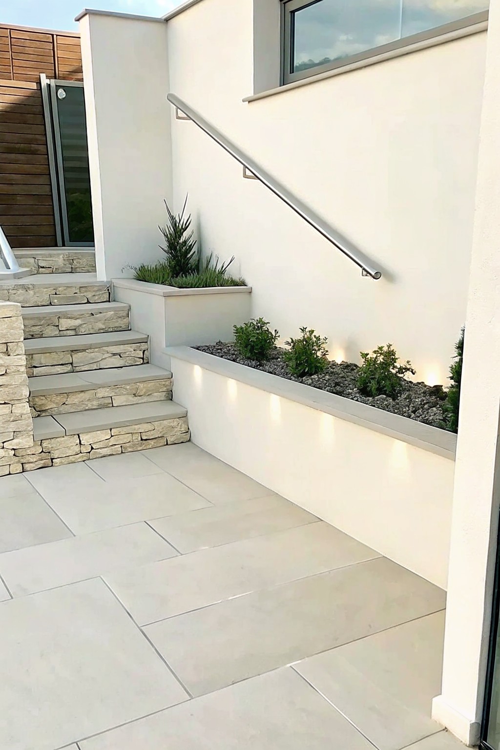 A modern outdoor patio area with light-colored tile flooring, stone steps, and a long raised concrete planter filled with gravel and small green plants, featuring integrated lighting along its front edge.