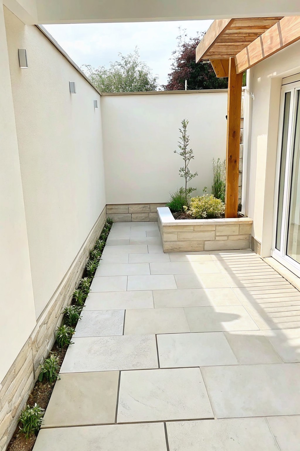A paved outdoor courtyard with a narrow border of low plants along the left wall and a raised stone planter bed on the right.