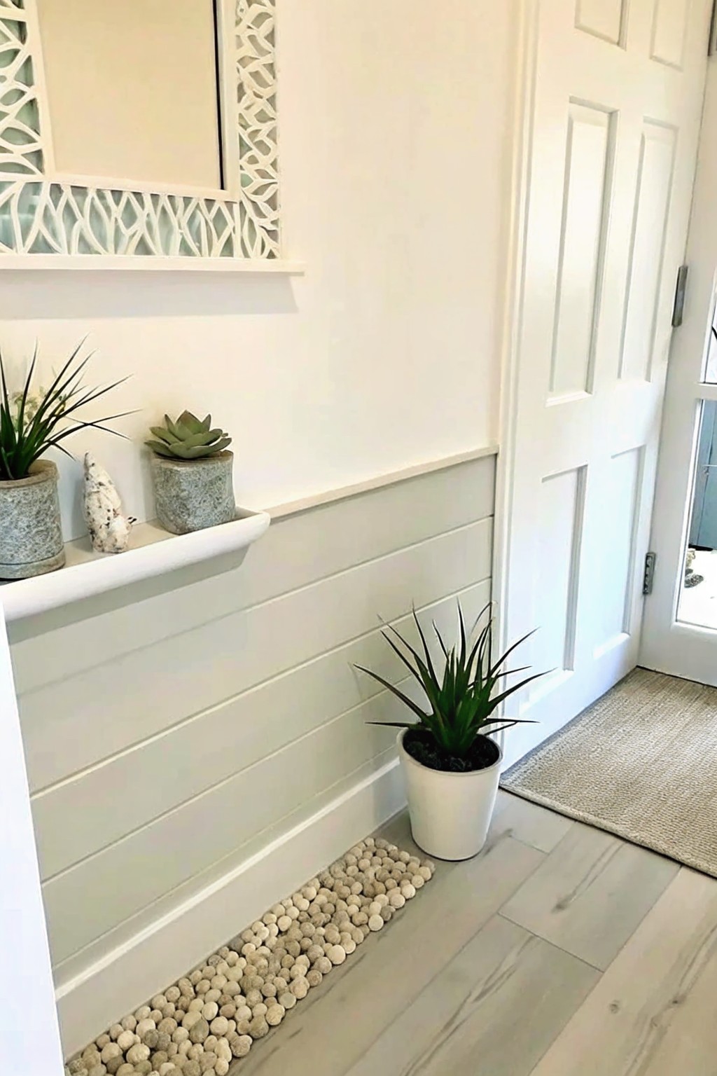 An interior entryway with white wainscoting, a floating shelf holding potted plants, a floor plant in a white pot, and a narrow border of small pebbles along the base of the wall next to light wood flooring.