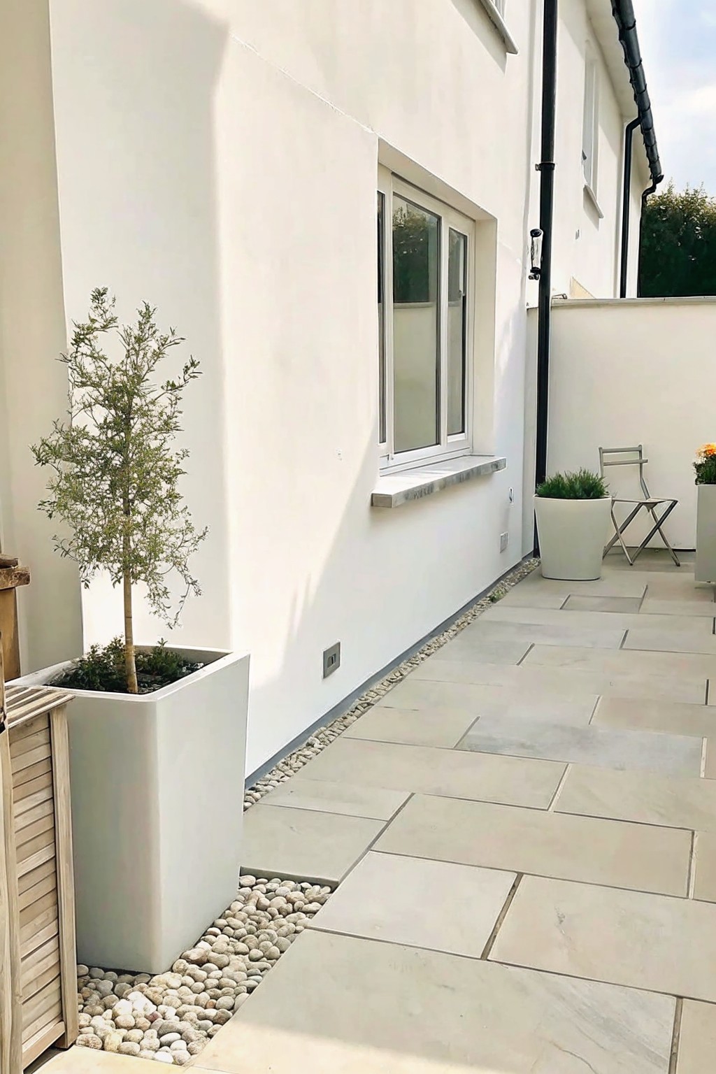 A paved outdoor area with large light stone slabs, a border of small pebbles along a white wall, and several white planters holding small trees and shrubs.
