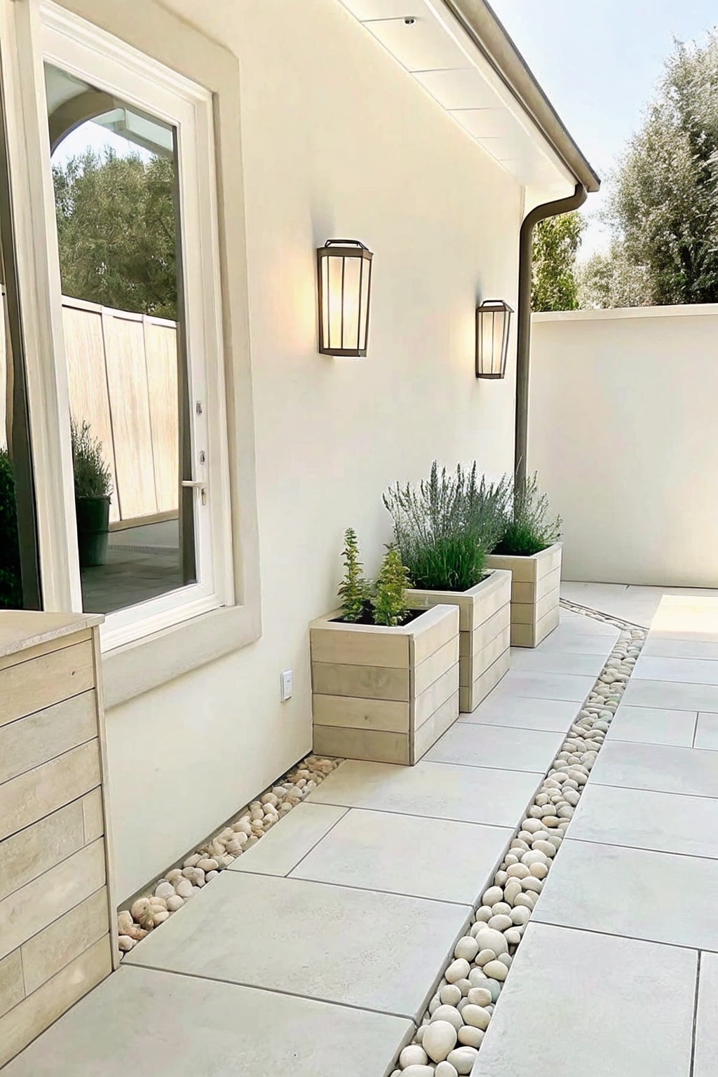 A light stone patio runs beside a house wall with a border of white pebbles separating it from three wooden planters holding shrubs.