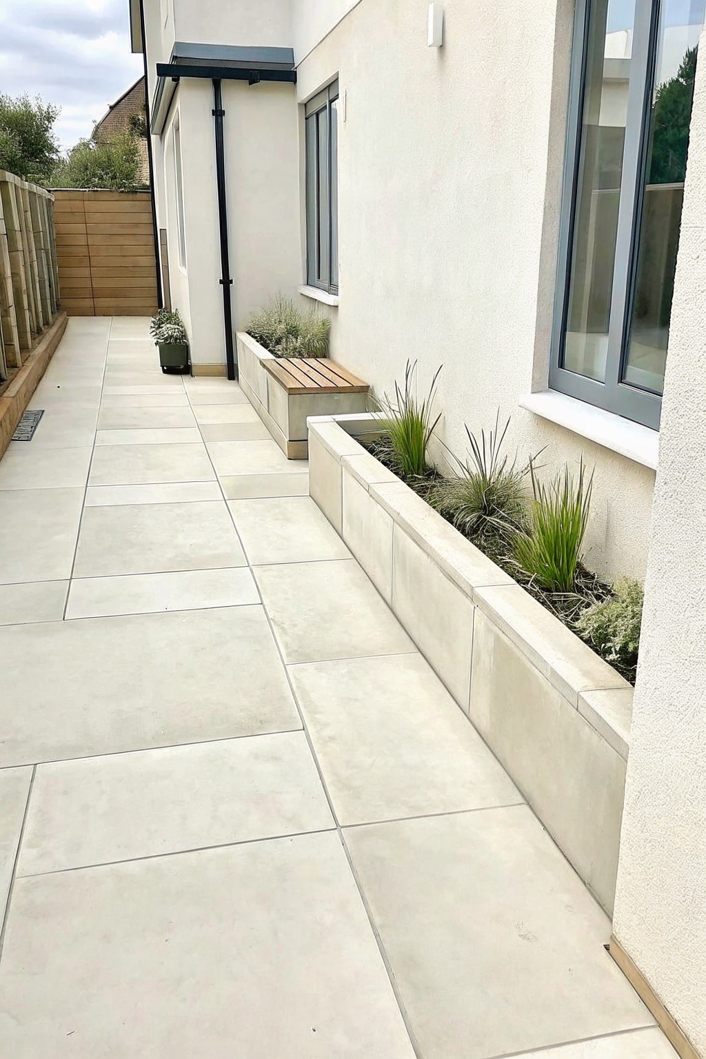 A modern house side path lined with long raised concrete planters containing layered grasses and small shrubs.