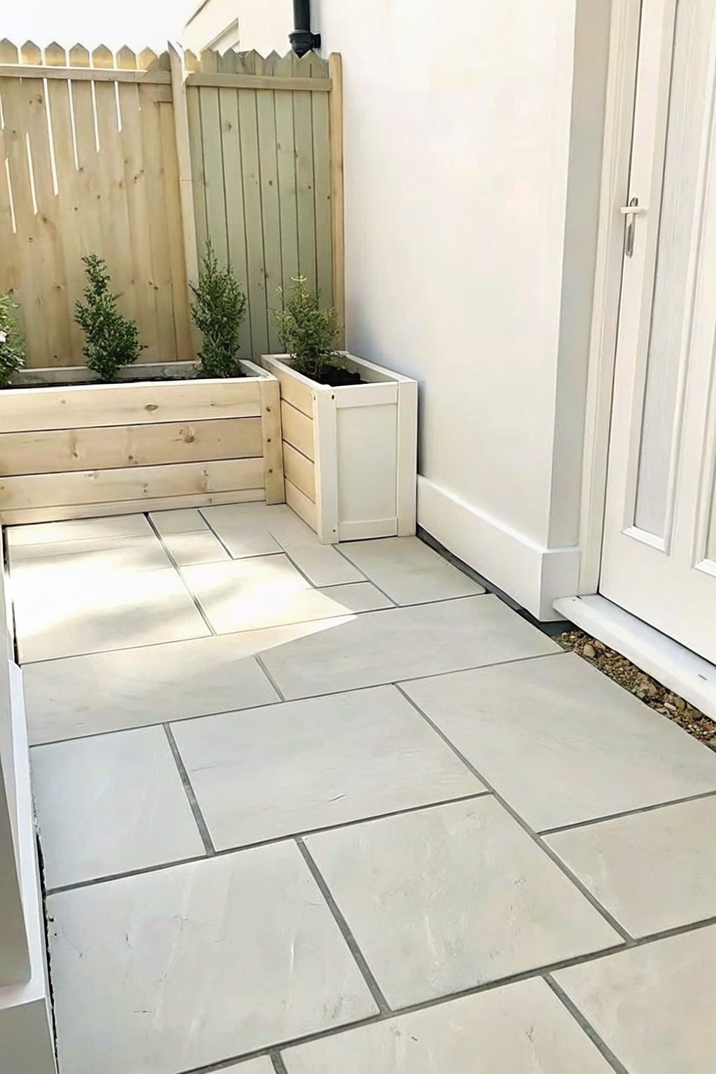 Paved patio with light tiles, wooden planters, shrubs, white door and fence