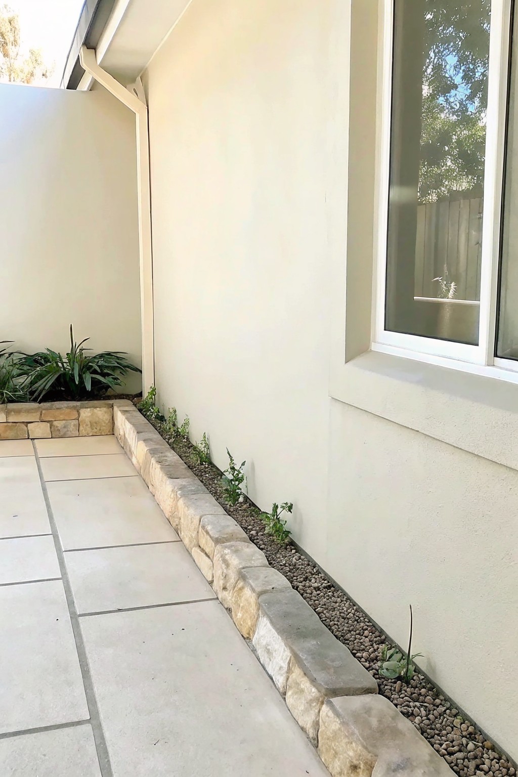 A narrow gravel bed with small plants is bordered by stacked stone along the base of a beige stucco house wall next to a tiled patio.