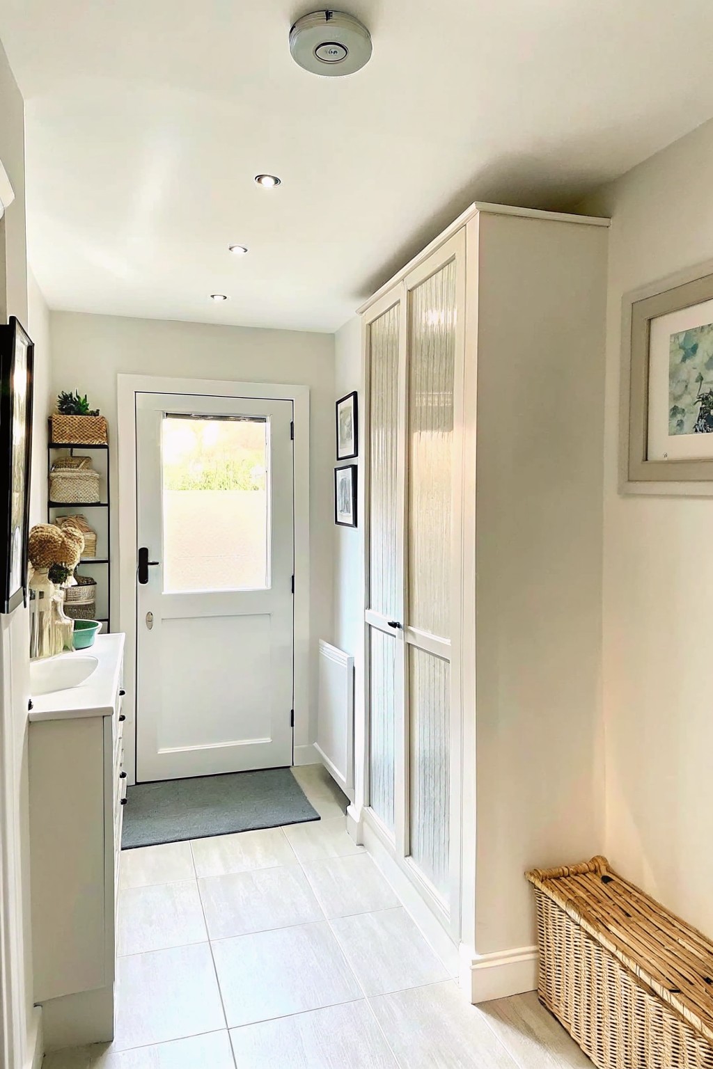 Narrow interior entry hallway with white walls, a white door at the far end, a tall white cabinet with ribbed glass doors along the right wall, and a white sink vanity on the left.