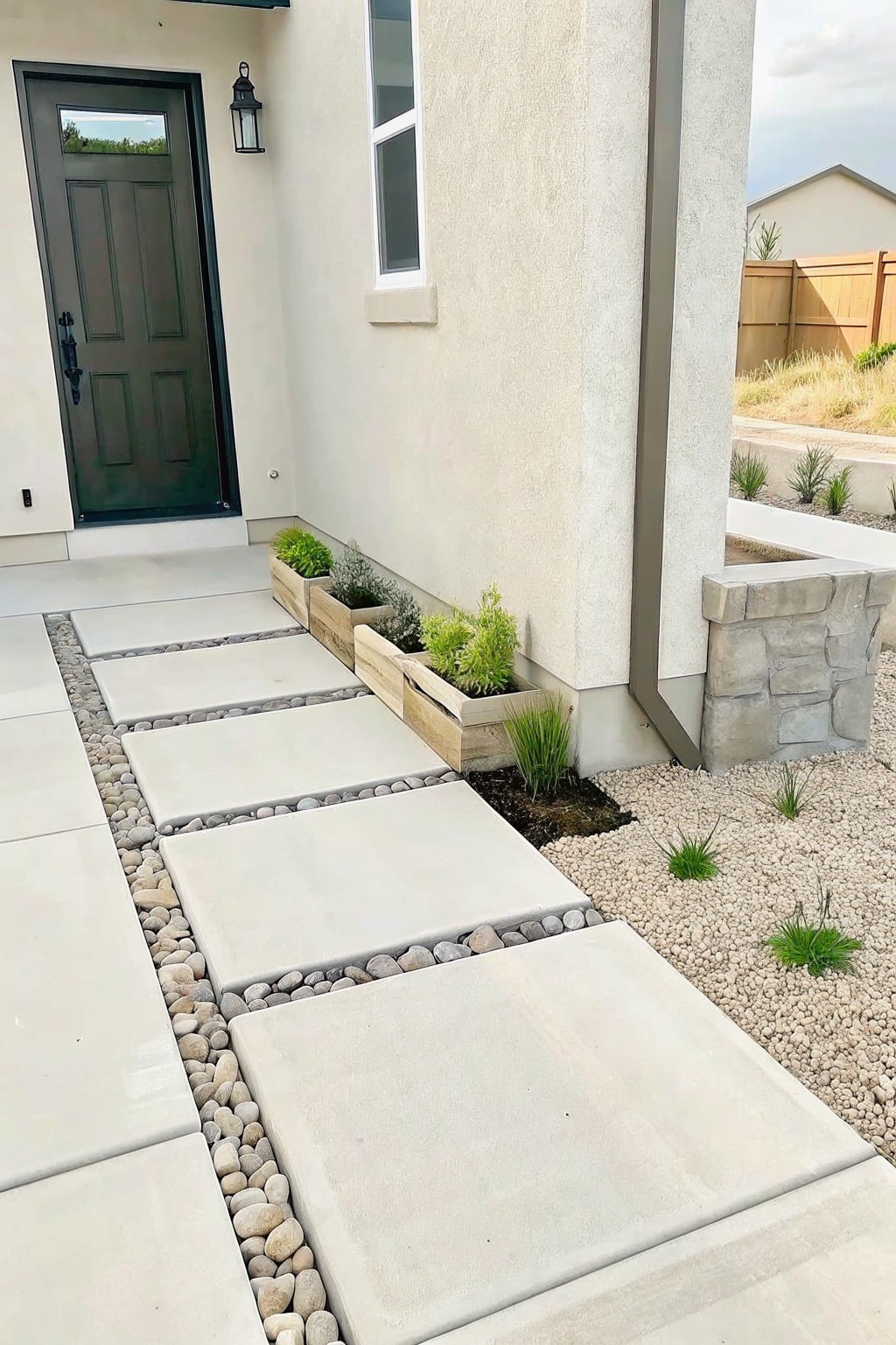Concrete paver path with pebble borders runs beside wooden planters filled with layered plants along a house exterior.