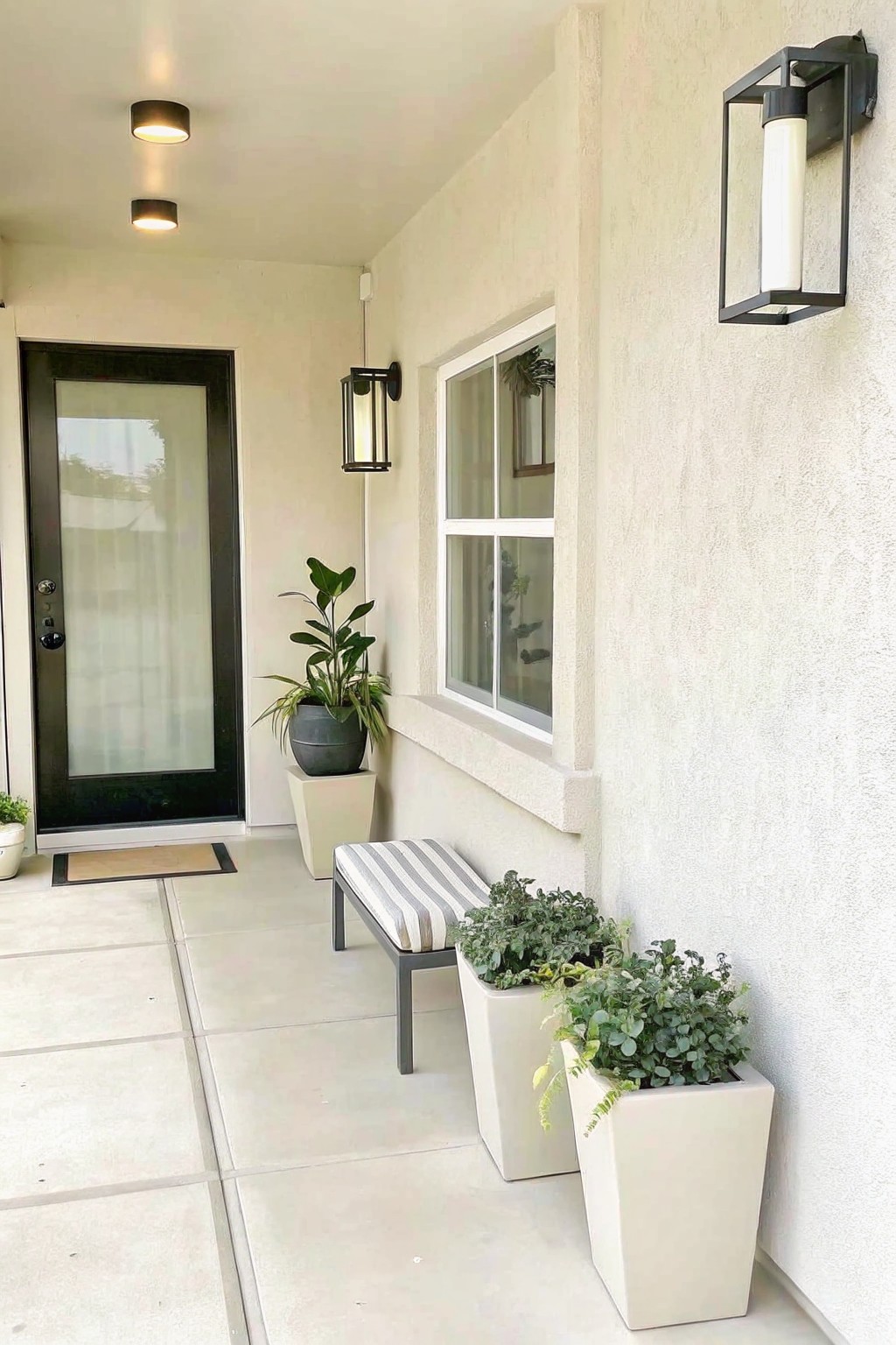 A covered house porch with a striped bench between two white planters filled with greenery, positioned next to a window and black door.