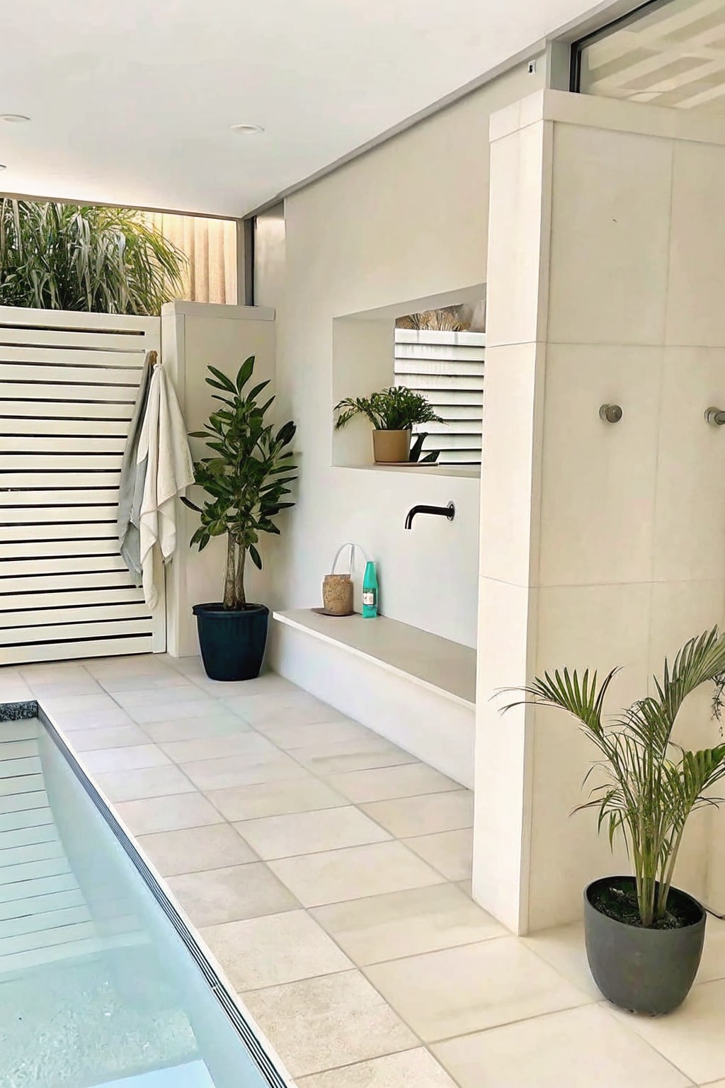 Outdoor poolside area with a long white built-in ledge under a black wall faucet, a recessed niche above it, potted plants on the ledge and floor, and a tiled deck.