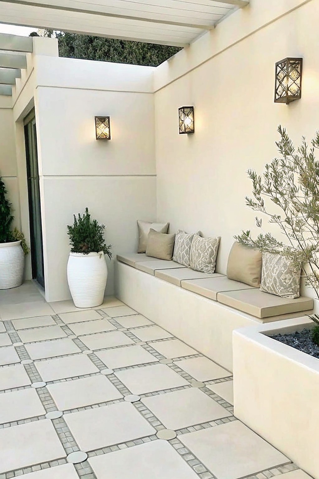 An outdoor patio with a long built-in bench along a white wall, beige cushions with patterned pillows, two large white planters, and wall sconces.