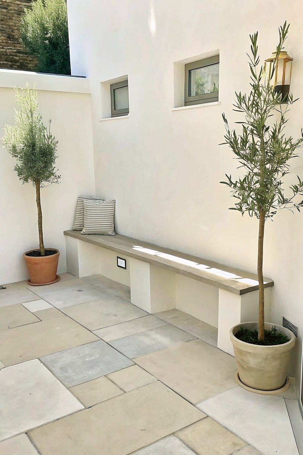 An outdoor patio with a long built-in bench along a white wall, two potted trees at each end, and large light-colored floor tiles.
