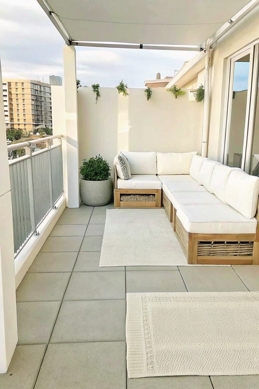 An L-shaped outdoor sectional sofa with white cushions and wooden base sits on a tiled balcony with two rugs and potted plants against a white wall.