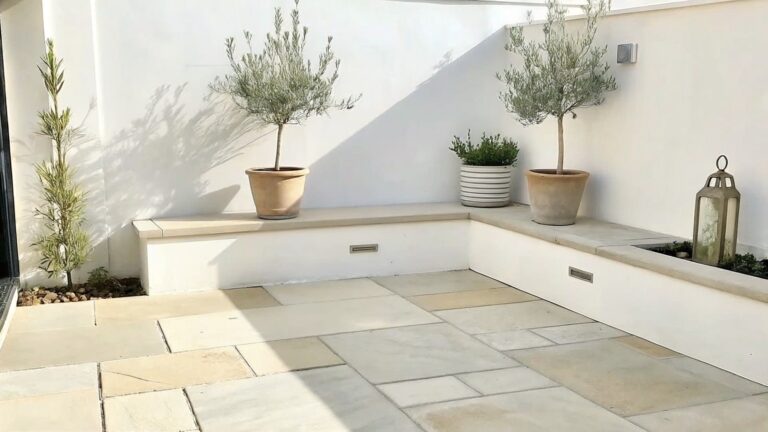 An outdoor patio with a long built-in bench along a white wall, two potted trees at each end, and large light-colored floor tiles.