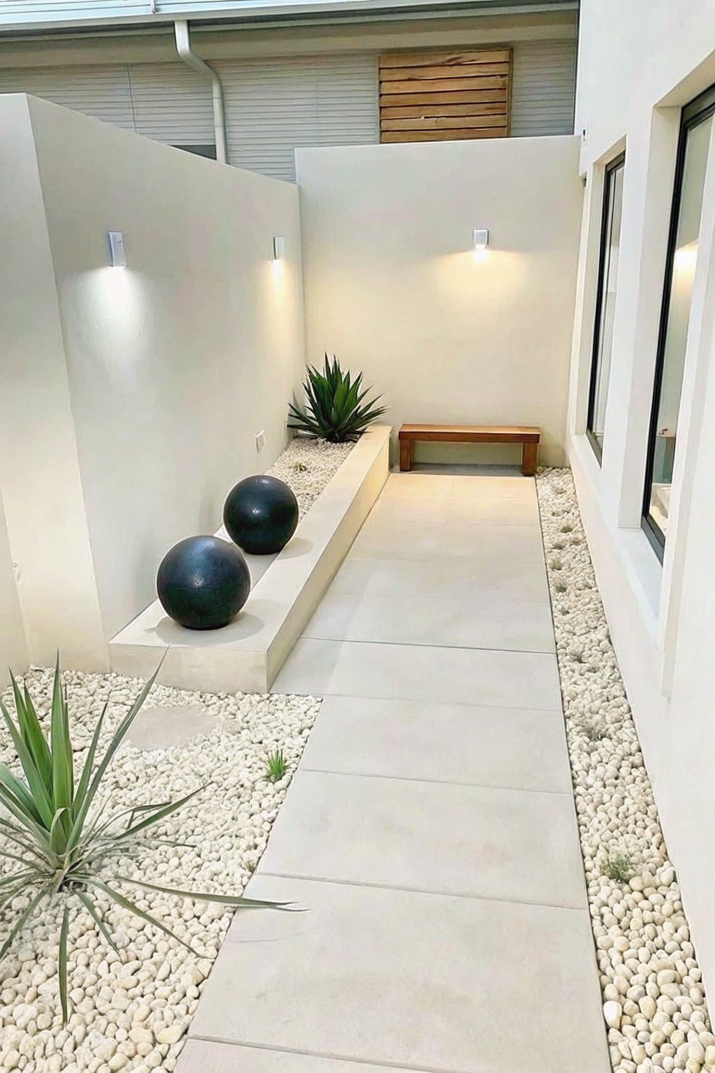 A narrow outdoor walkway of large light pavers runs between white gravel beds holding two dark spheres, a raised planter with an agave, and a wooden bench at the far end.