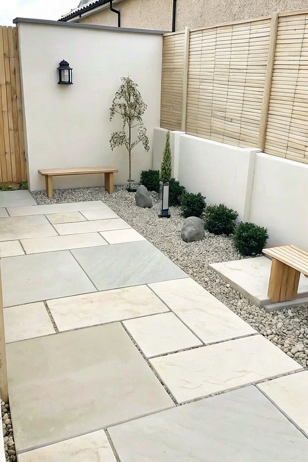 A backyard garden features large light stone pavers forming a path beside a gravel bed with small shrubs, rocks, and a young tree, set against white walls and wooden fencing.