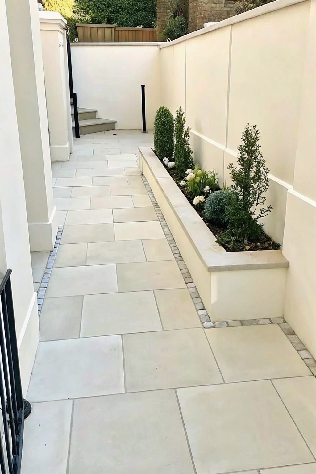 Narrow stone walkway beside raised planter with green shrubs against beige walls.