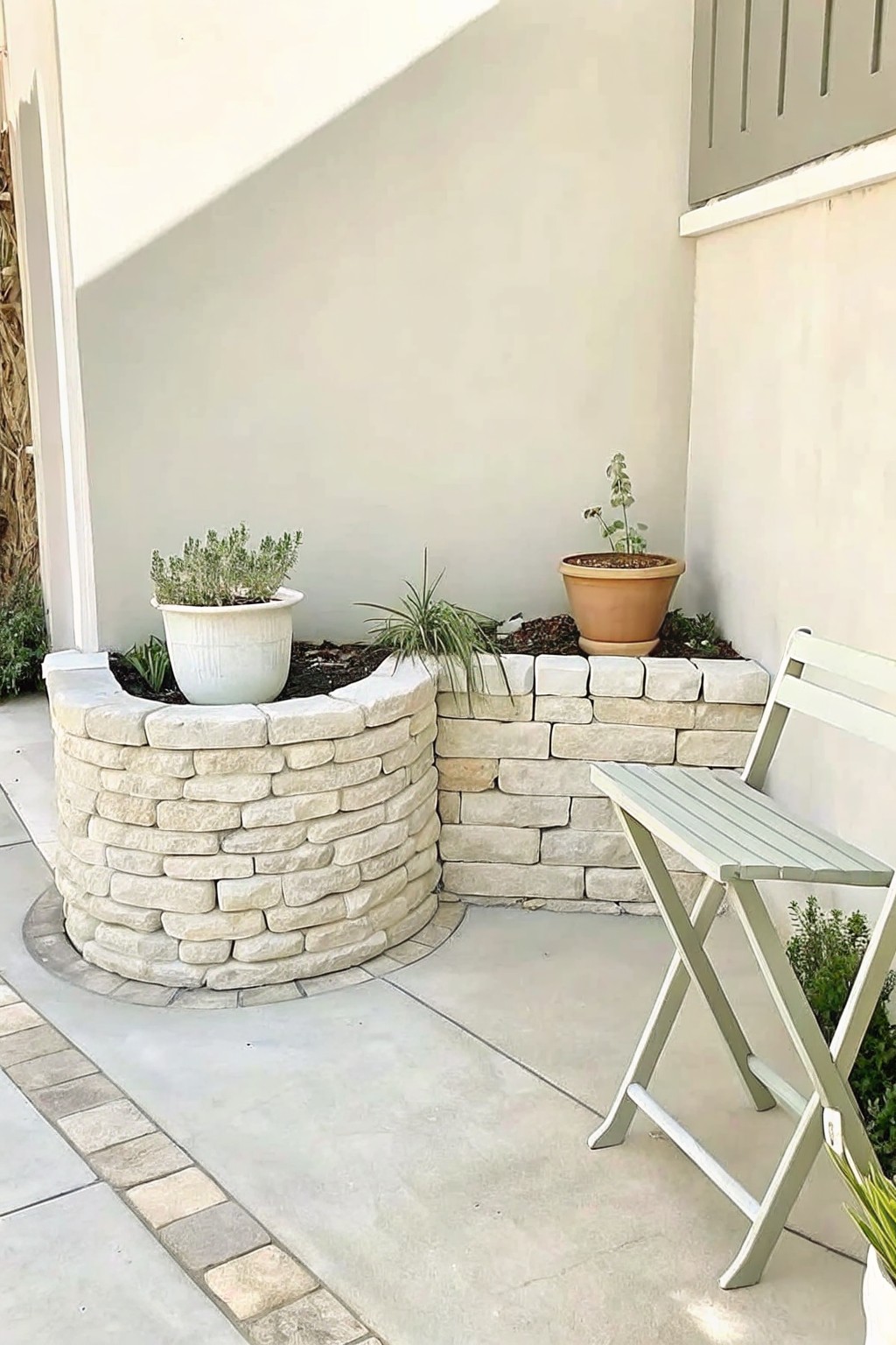 Curved stone planter with potted plants and green folding chair on patio.