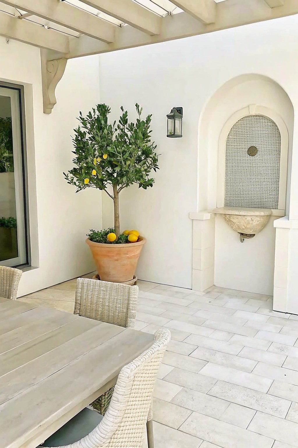 White-walled patio with lemon tree in pot, arched mosaic fountain, and woven chairs