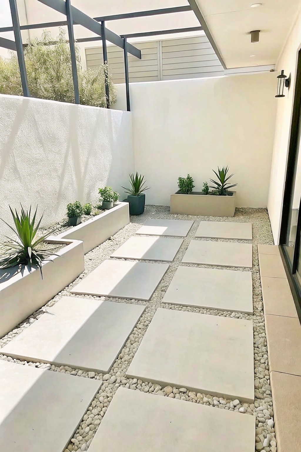 A narrow outdoor courtyard with large square light-colored pavers set into white gravel, bordered by raised concrete planters holding small green plants.