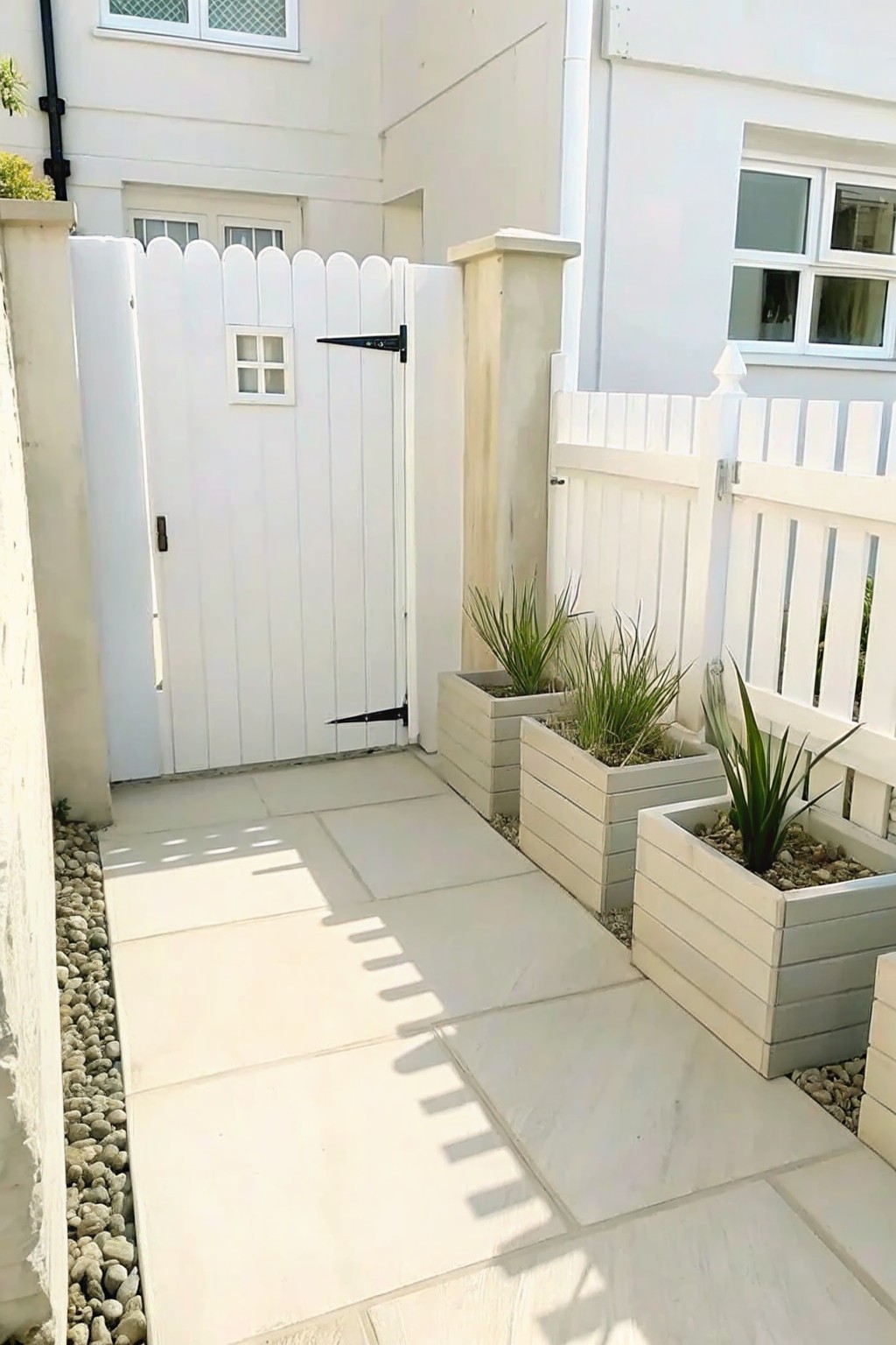 A stone pathway runs beside several light-colored raised planters with green plants, next to a white fence and gate on a house exterior.