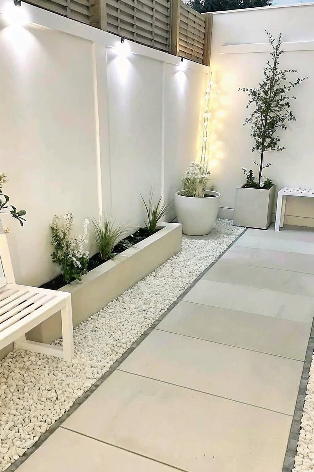 Modern courtyard walkway with white gravel, planters, bench, potted plants, and string lights.