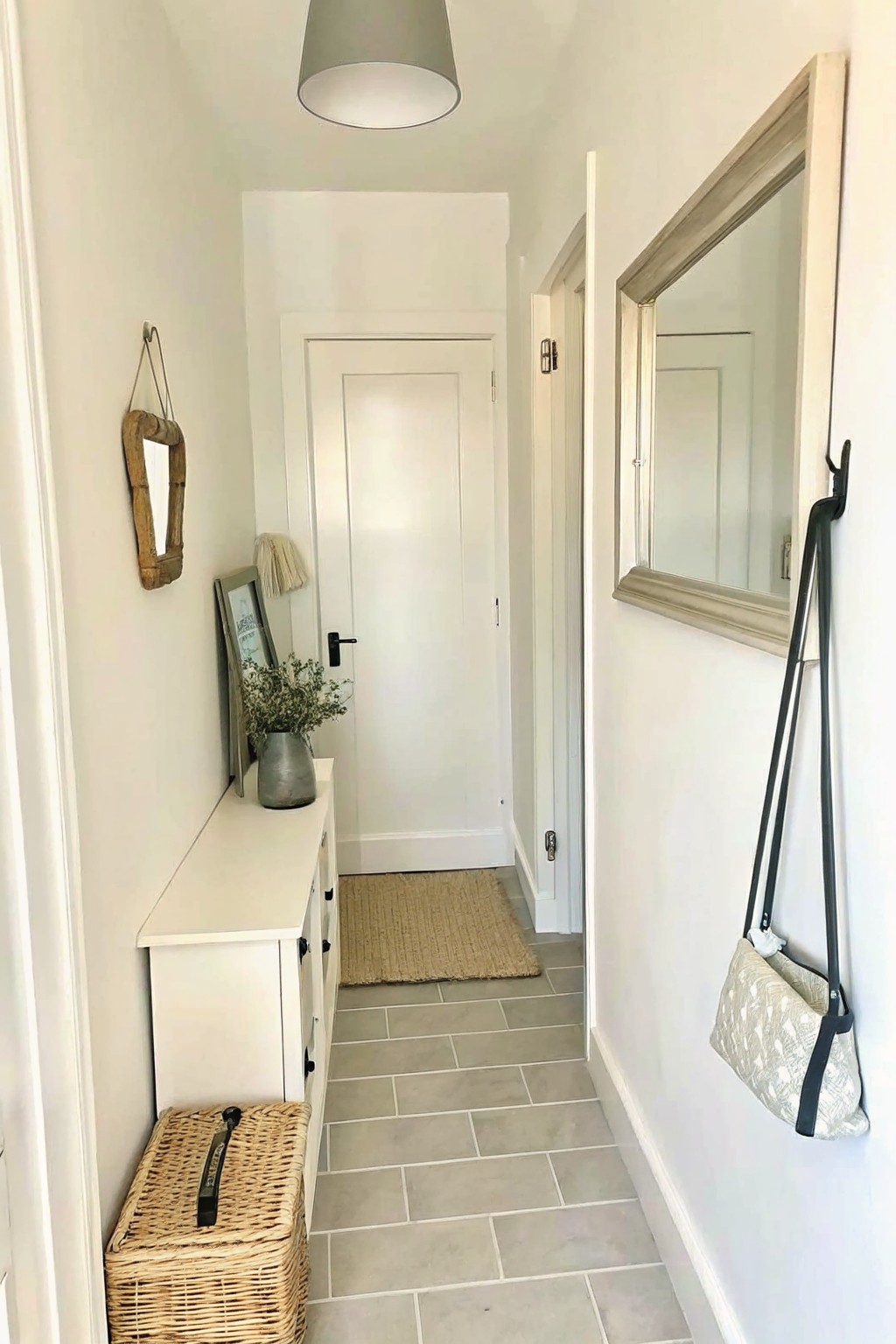 A narrow white hallway features a low console table with a basket underneath, a small mirror on the left wall, and a patterned bag hanging on the right wall, with a door visible at the far end.