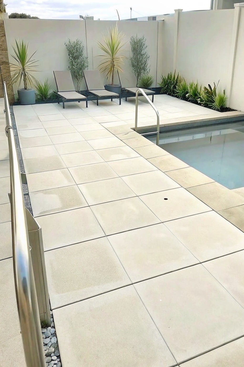Tiled pool deck with two lounge chairs, potted plants, and stainless steel handrails