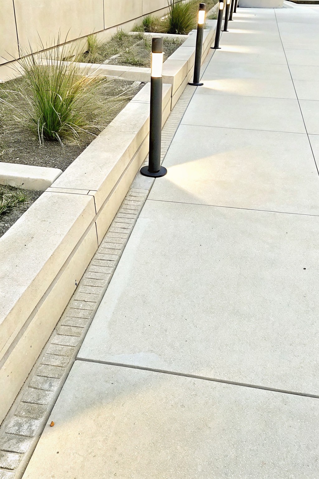 Modern concrete walkway lined with glowing black bollard lights beside grassy planter.